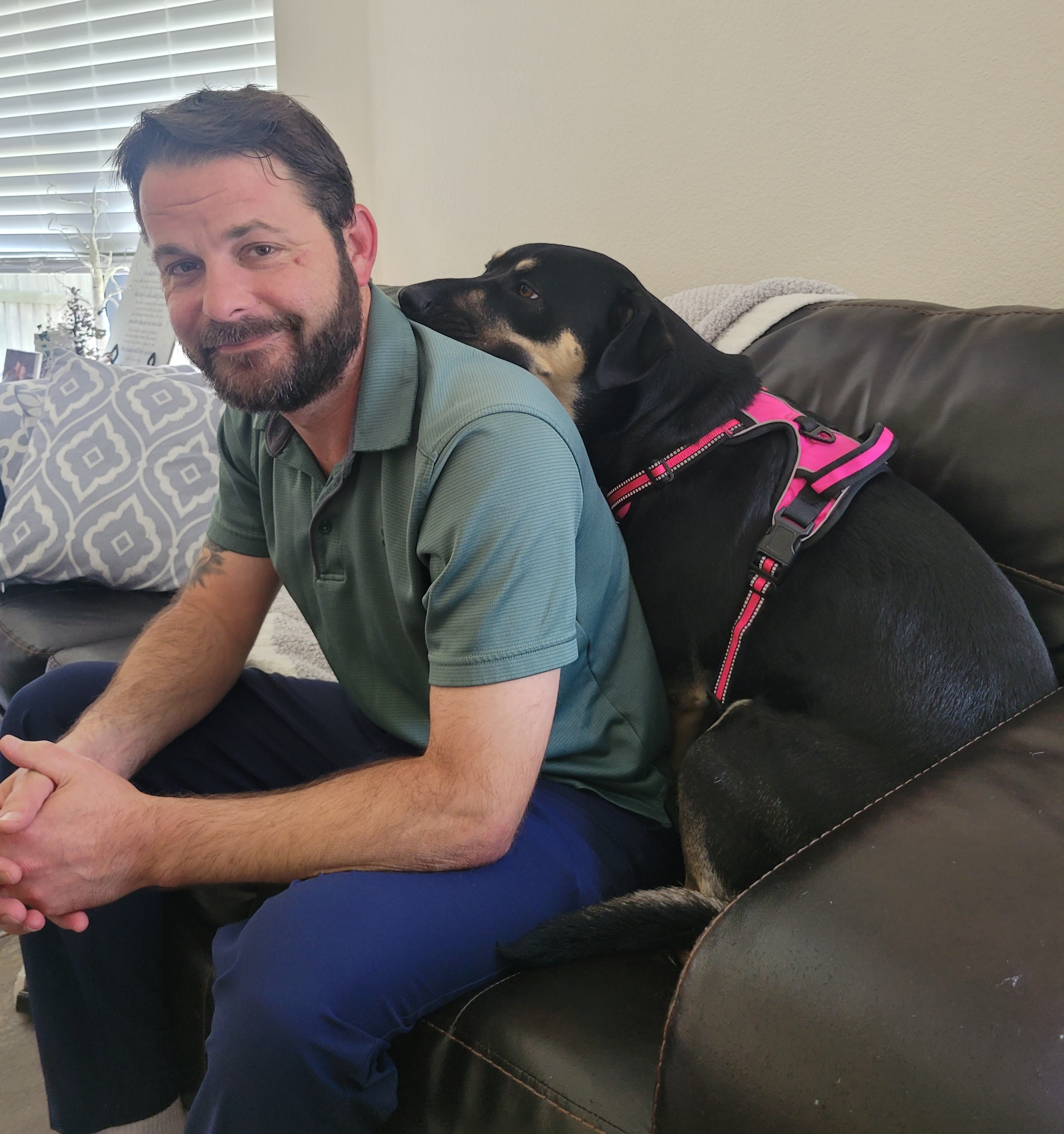 Kona, a large black and tan dog, sitting behind her adoptive dad on a brown leather sofa. Kona is resting her head on his back.