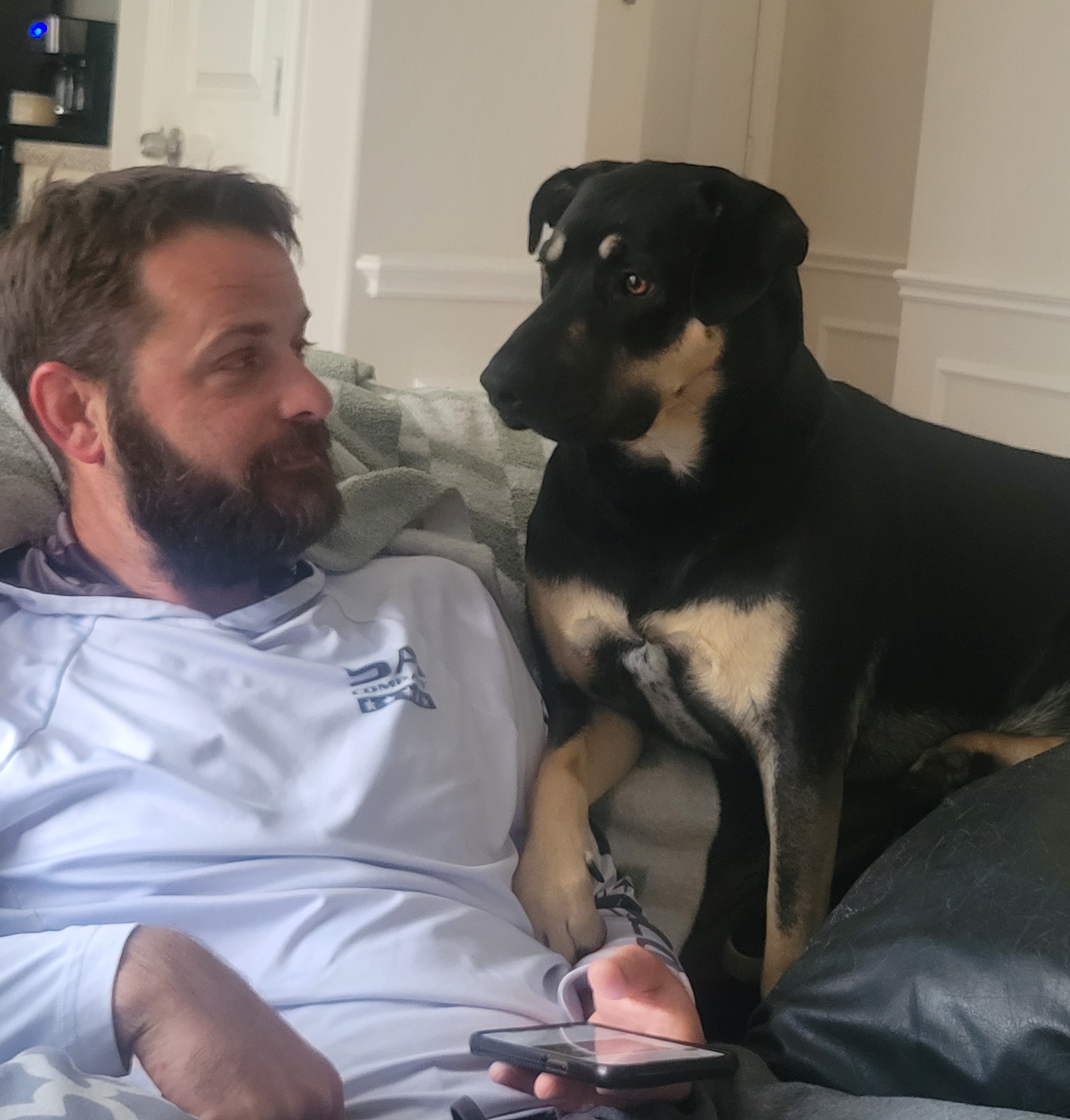 Kona, a large black and tan dog, sitting on a sofa next to her adoptive dad. They're looking at each other. Dad is smiling at Kona.