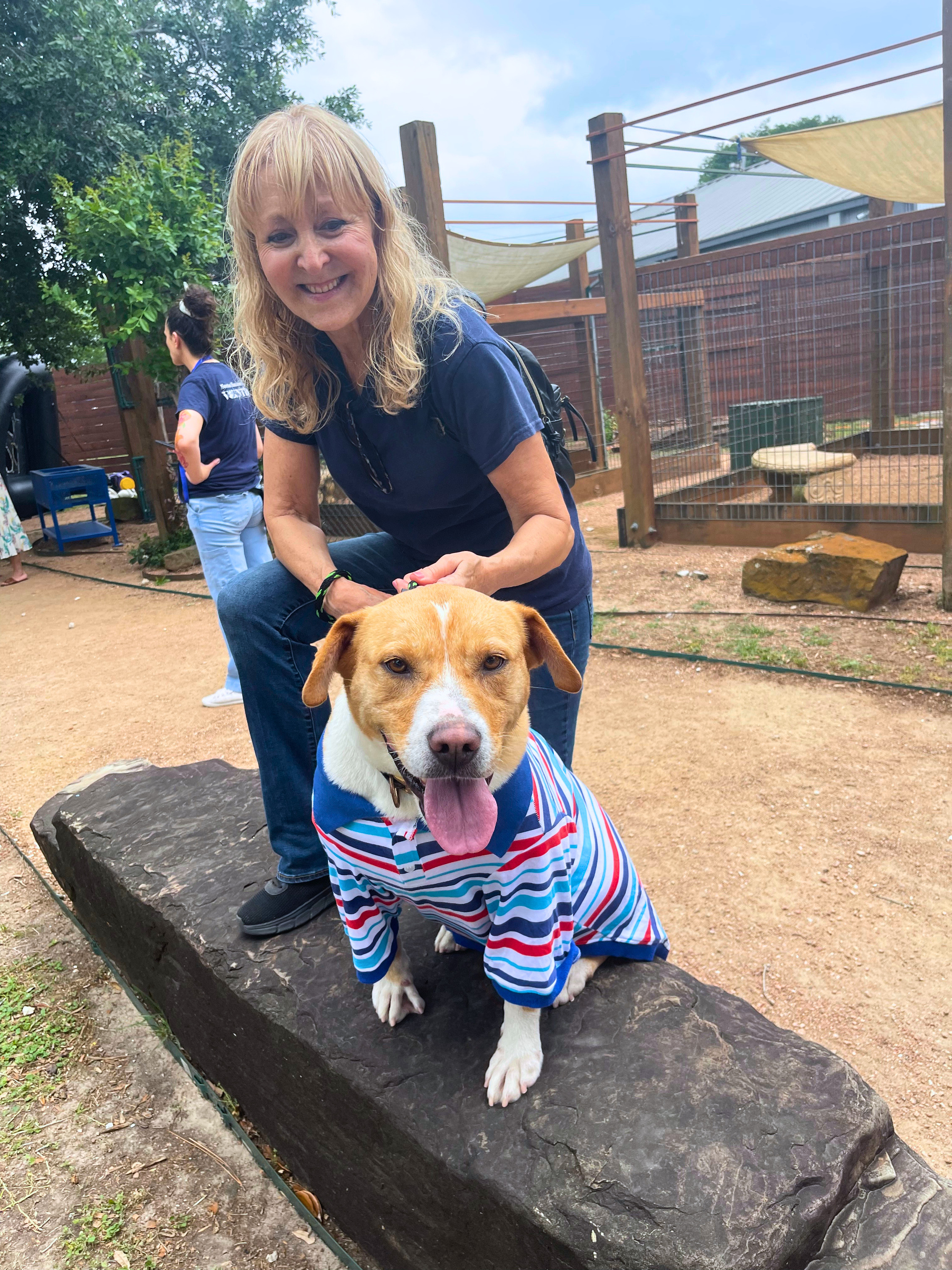 Outdoors; Brigitte, a volunteer, stands behind a dog who is wearing a striped polo shirt.