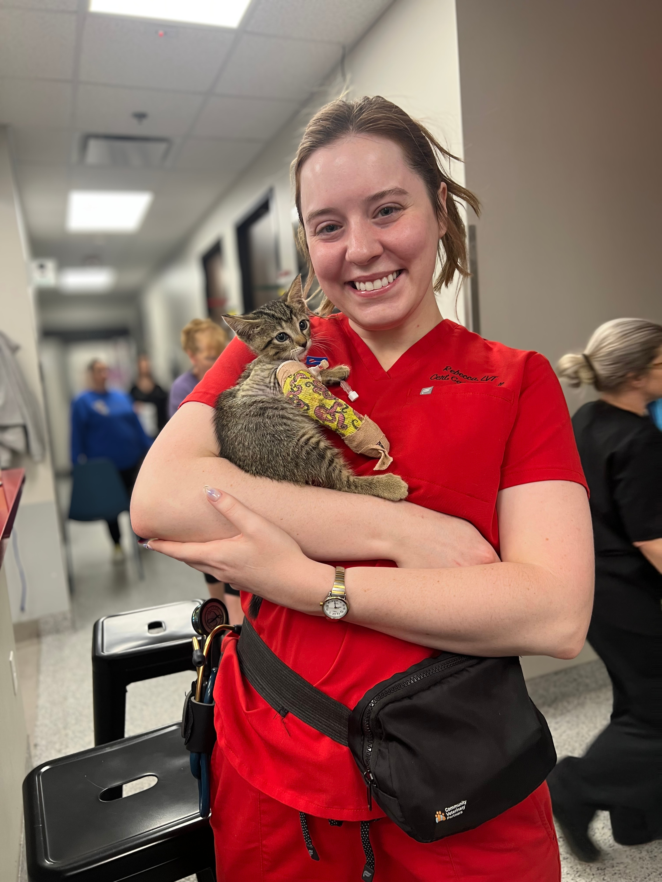 A smiling woman in red scrubs holding a kitten. The kitten has a yellow cast on its front leg.